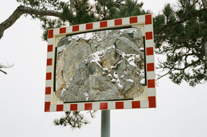 A mirror mingling with snowy pine branches. Around its face the red and white checker of caution, and in the center a stoney reflection of the rocks behind.