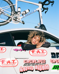 Racing Dream Team member Niara leans out the window of the 911 Dakar team car from FAT International, with lots of Dream Beyond graphics and a Specialized SL8 on the roof rack.