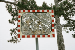 Alternate frame of a mirror mingling with snowy pine branches. Around its face the red and white checker of caution, and in the center a stoney reflection of the rocks behind.