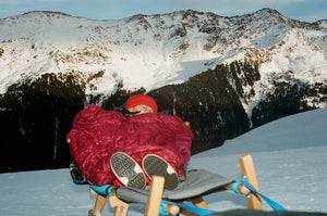 A snowy mountain summit, in the corner a child bundled in striking red.